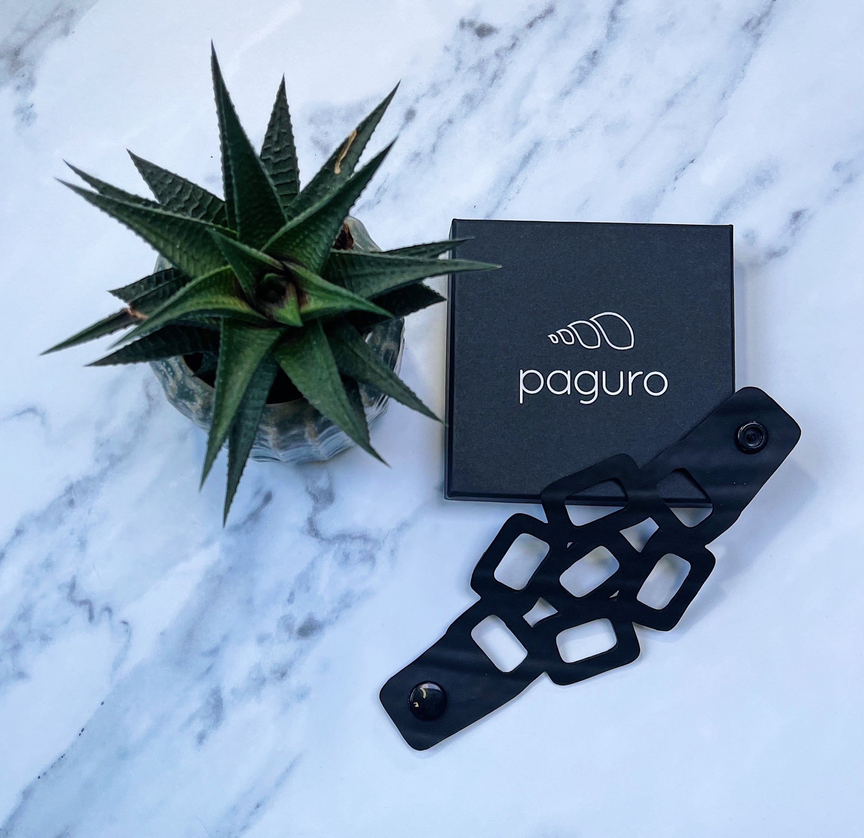 Paguro Upcycle eco friendly chain bracelet in black displayed next to plant and branded box on marble surface